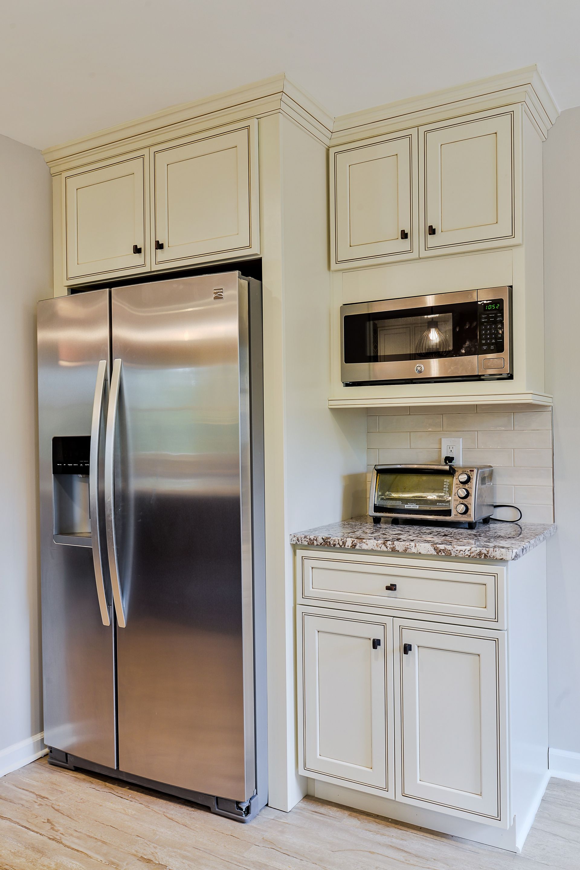 A stainless steel refrigerator next to cabinets with a microwave, toaster, and granite countertop in a light-toned kitchen.