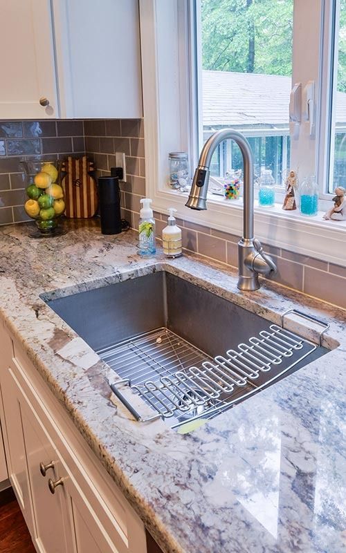 A kitchen sink with a metal faucet, a drying rack, and a granite countertop, with a fruit bowl and window in the background.
