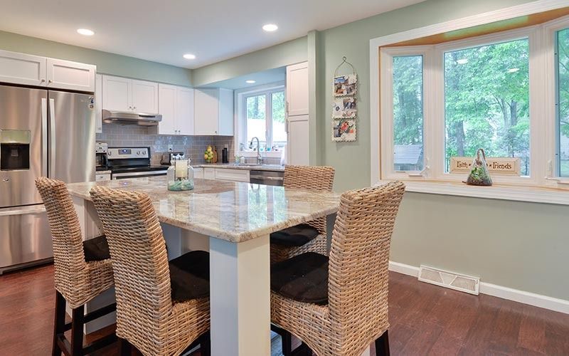 A kitchen interior with a marble-topped island, four wicker chairs, white cabinetry, and a large bay window.
