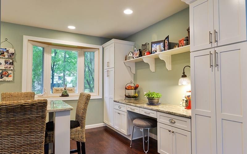 A light green dining nook with a white built-in desk, shelving, cabinets, and a wicker chair by a bay window.