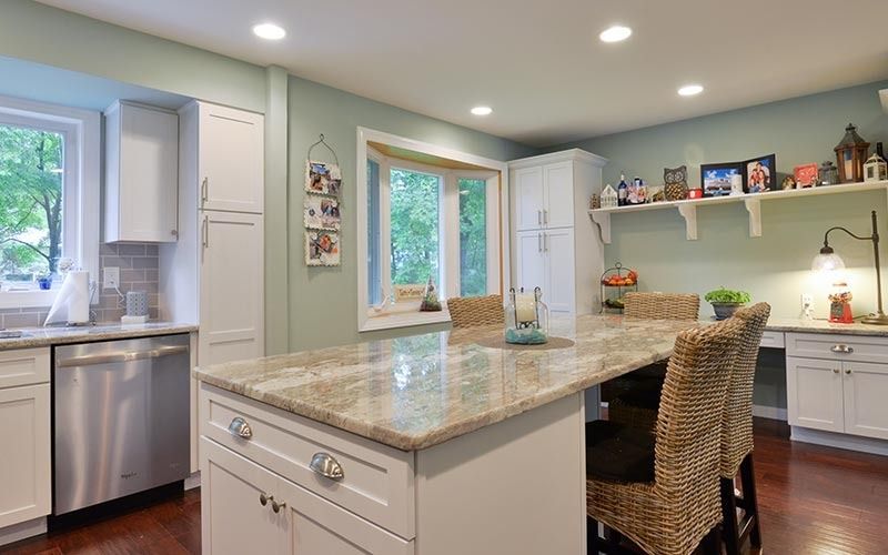 A bright kitchen with white cabinets, light green walls, a large granite island, and two woven chairs.