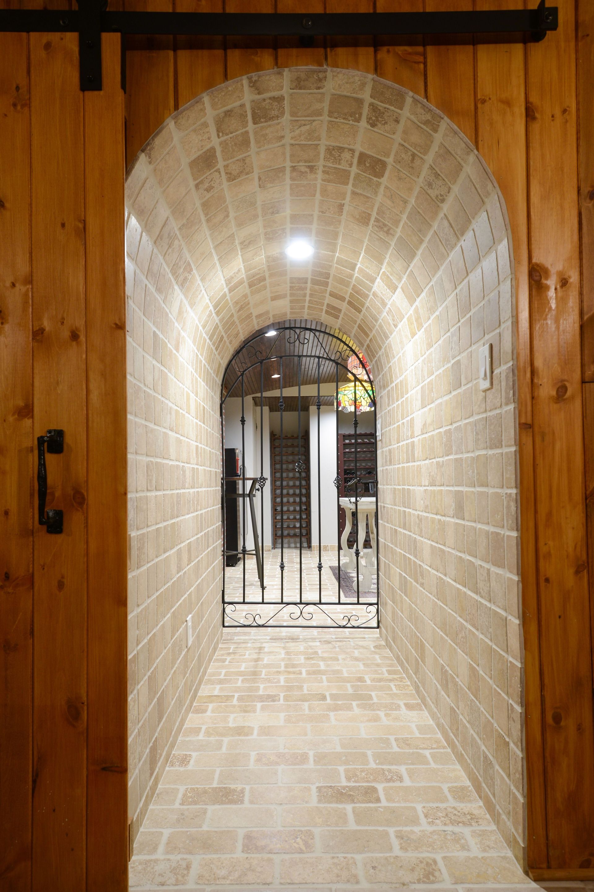 A vaulted brick hallway leads through a dark decorative gate, framed by wood paneling and a sliding barn-style door.