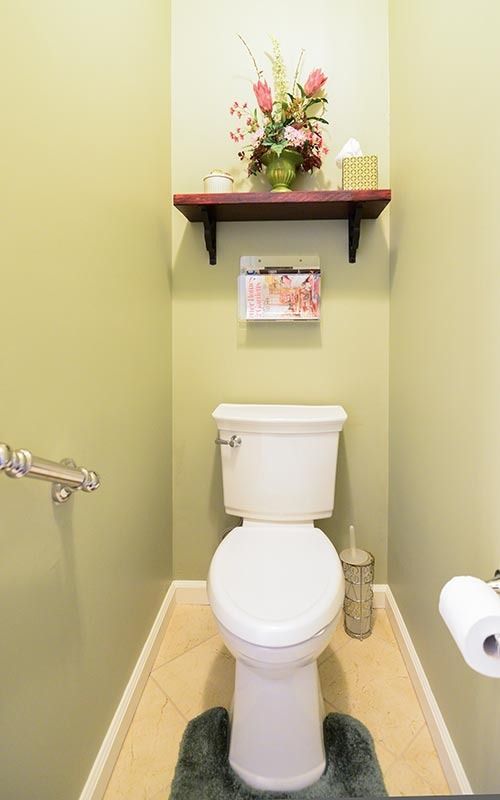 A small powder room with pale green walls, a white toilet, a brown floating shelf with flowers, and a toilet paper holder.
