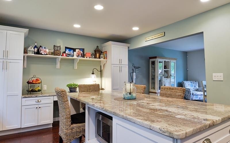 A bright kitchen featuring a large stone-topped island with wicker chairs, white cabinetry, and an open entryway.