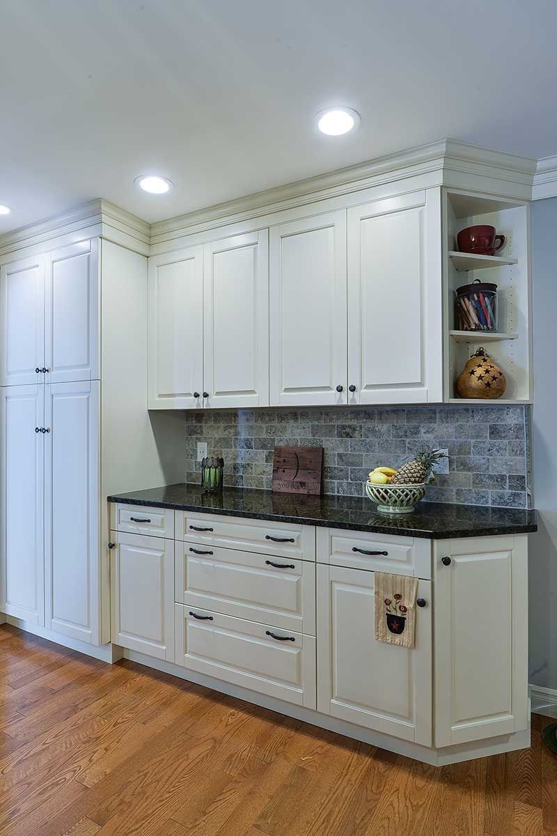 Off-white kitchen cabinets with black countertops and a rustic brick backsplash, featuring an open shelving unit.