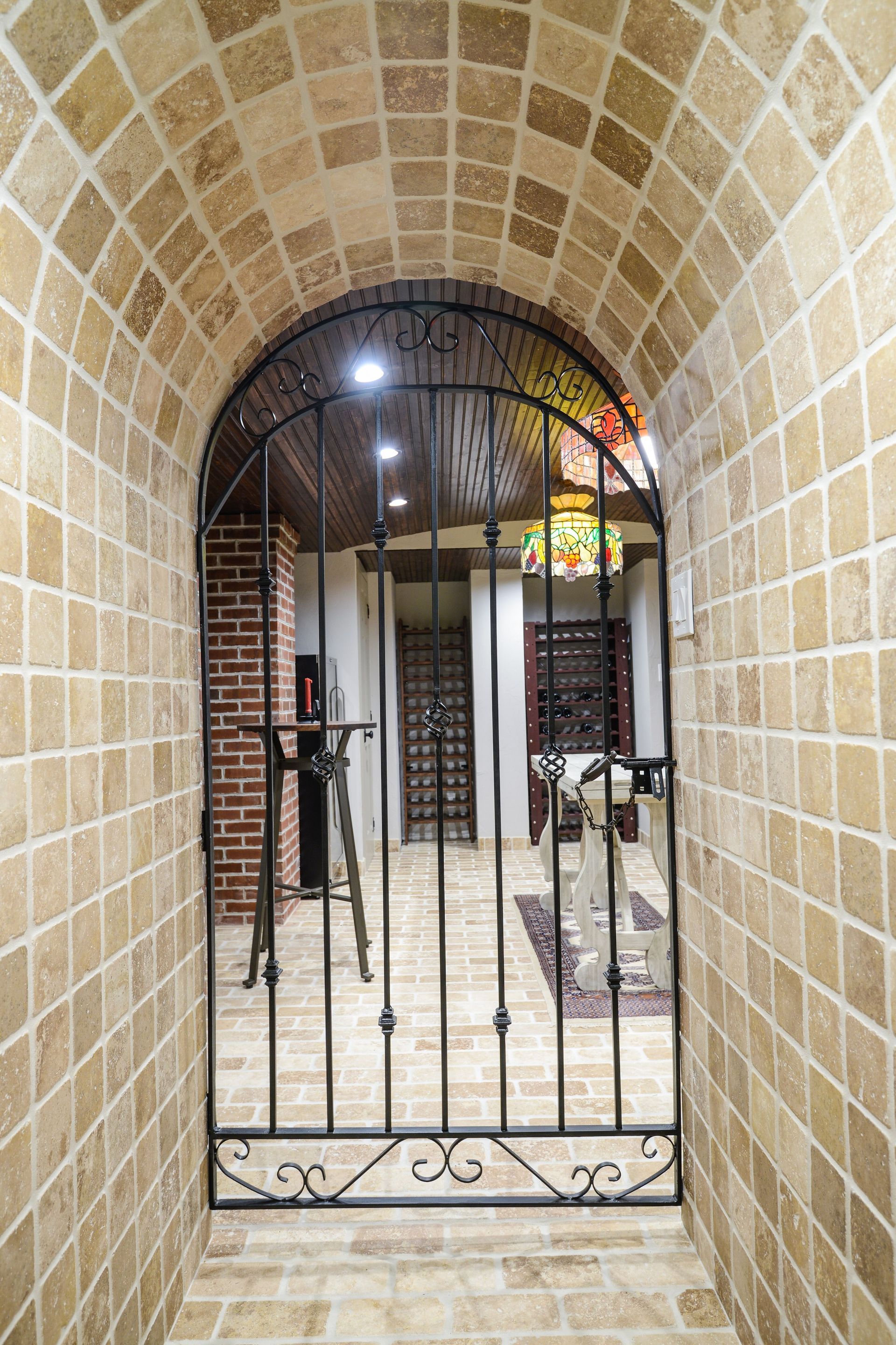 A stone arched hallway leads through a black wrought-iron gate into a wine cellar with brick accents and a stained lamp.