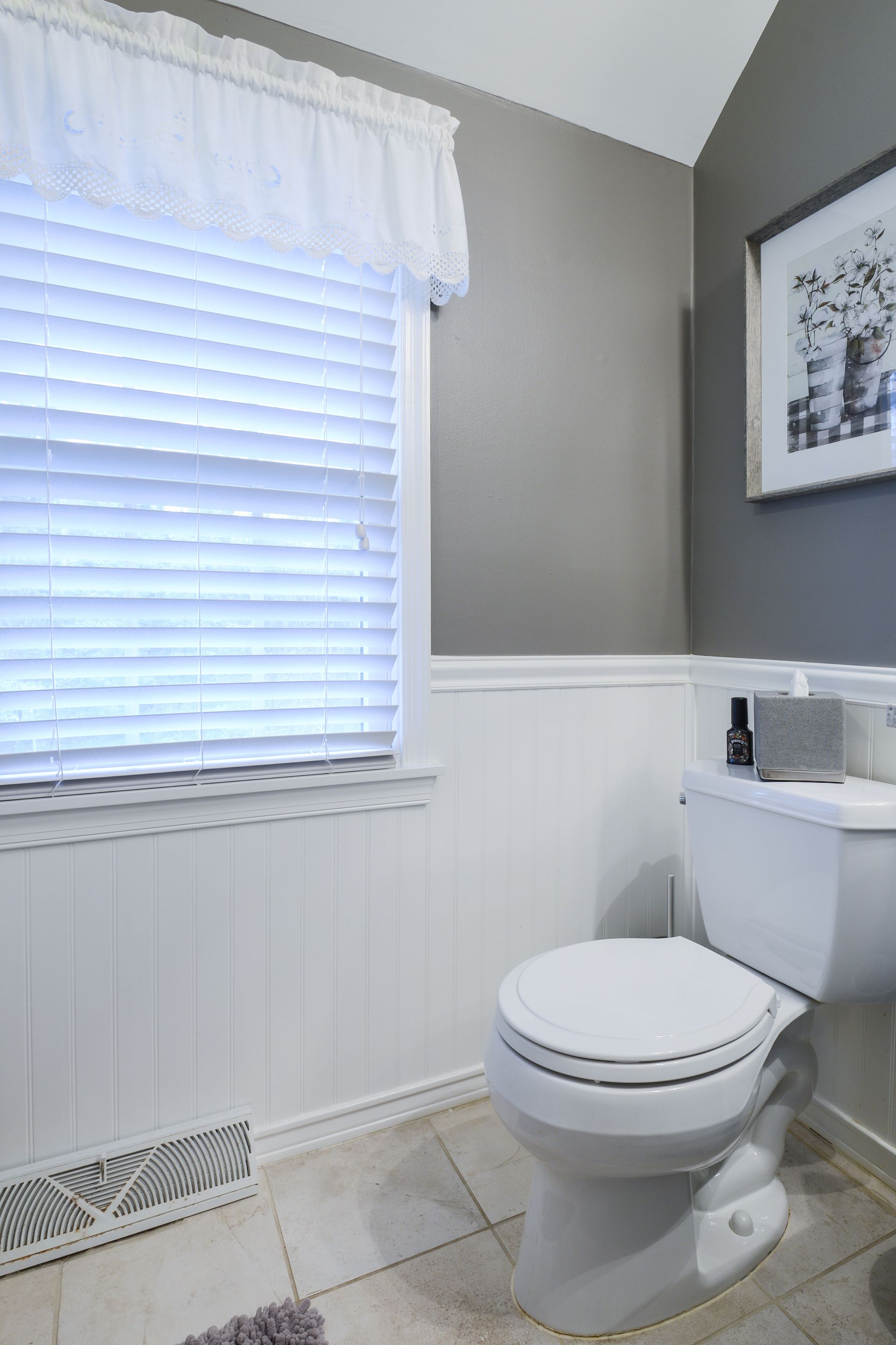A bathroom corner with a white toilet against a two-tone wall, featuring a window with white blinds and framed wall art.