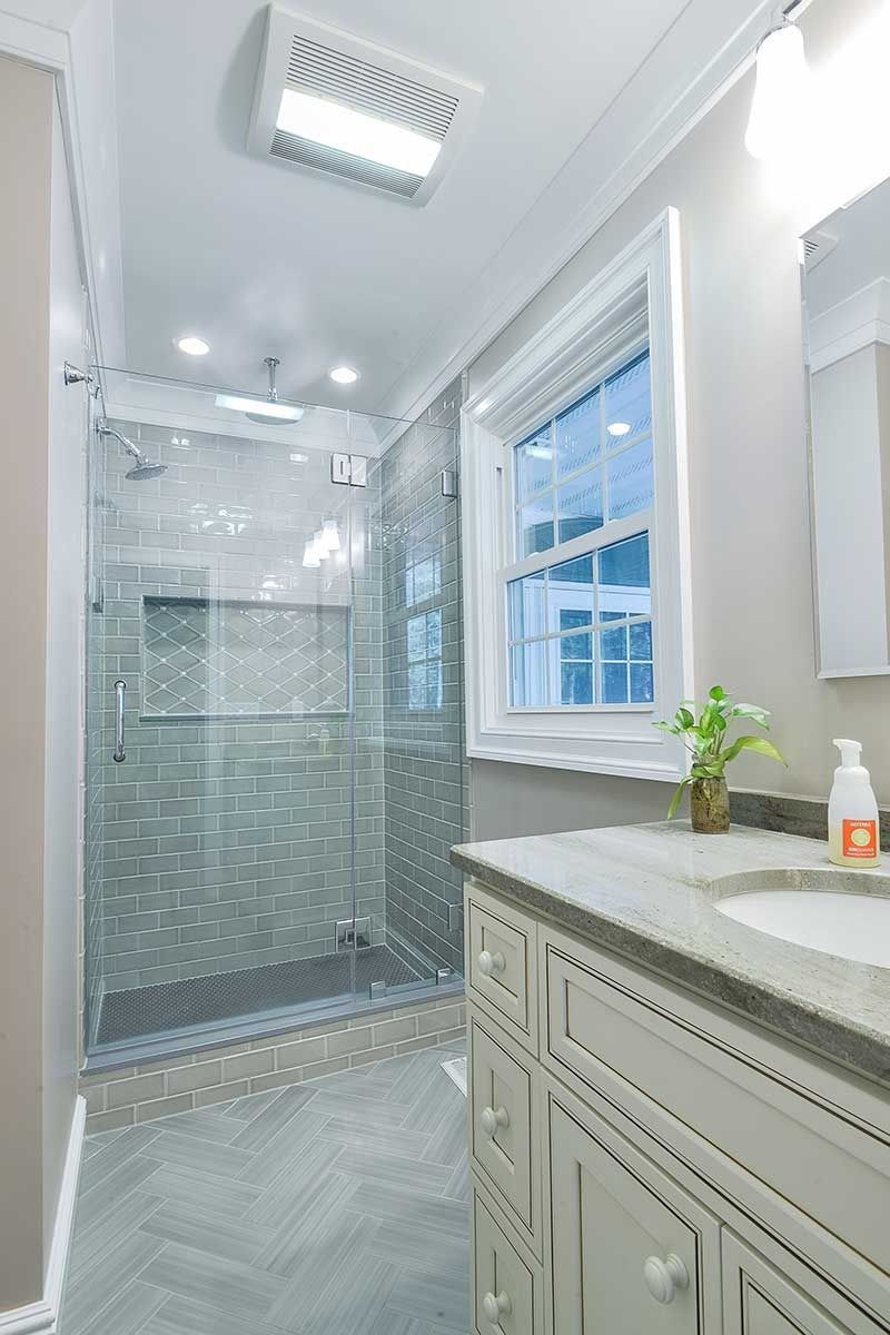 A modern bathroom featuring a glass-enclosed shower with gray tiled walls, a herringbone floor, and a white vanity cabinet.