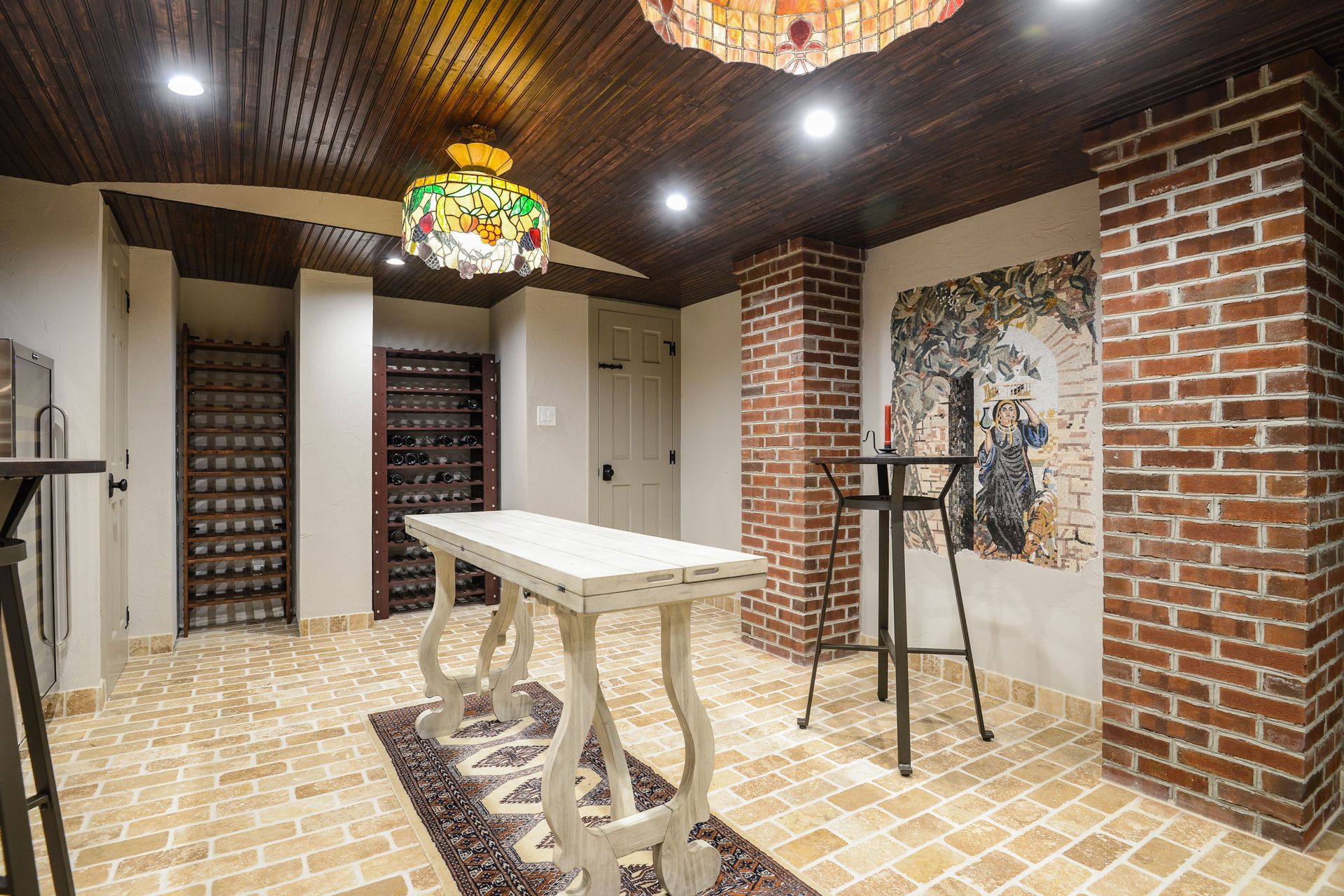 A wine cellar with brick columns, tiled floor, a white table, tall stools, wine racks, and stained-glass lighting.