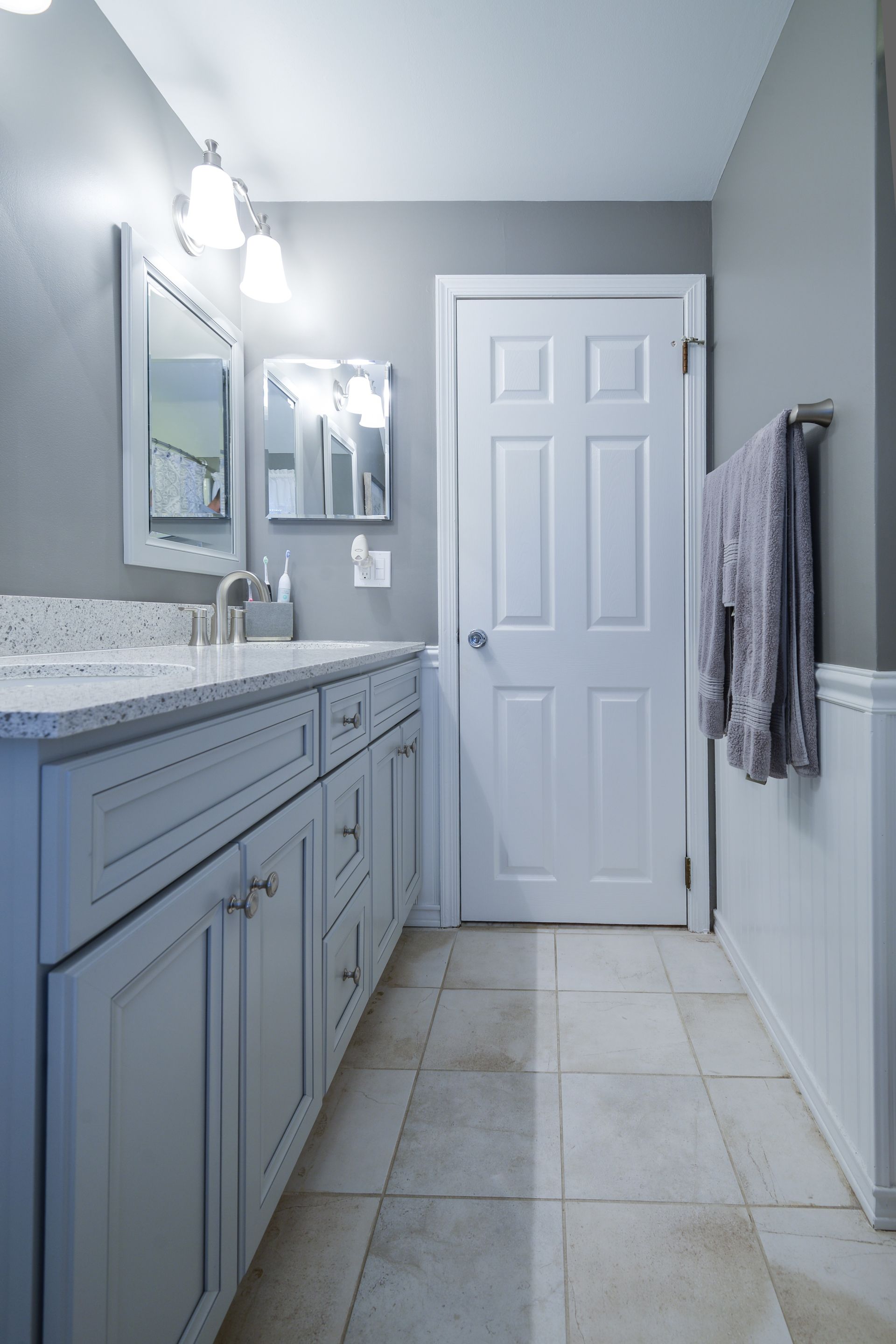 A modern bathroom featuring gray cabinets, light granite countertops, a white door, and tile flooring.