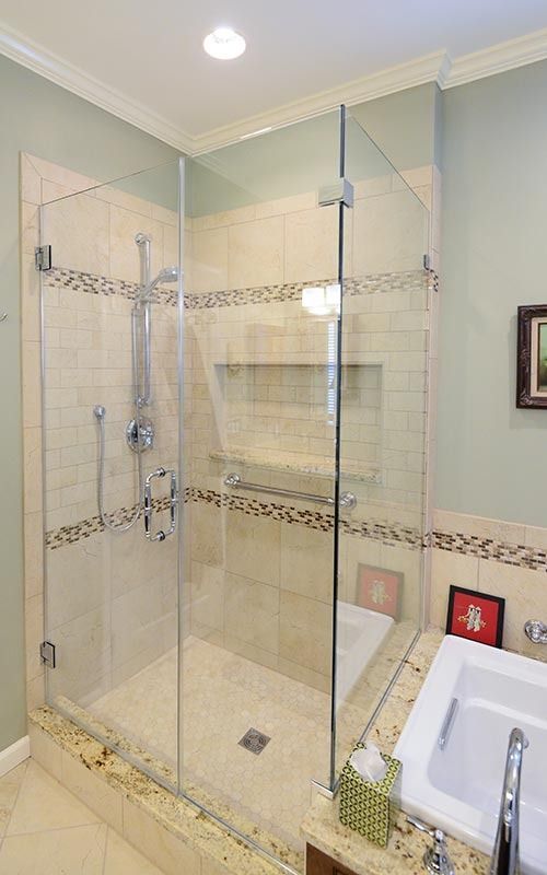 Modern glass shower stall with light beige tile, a patterned accent band, and built-in shelving, next to a bathtub.