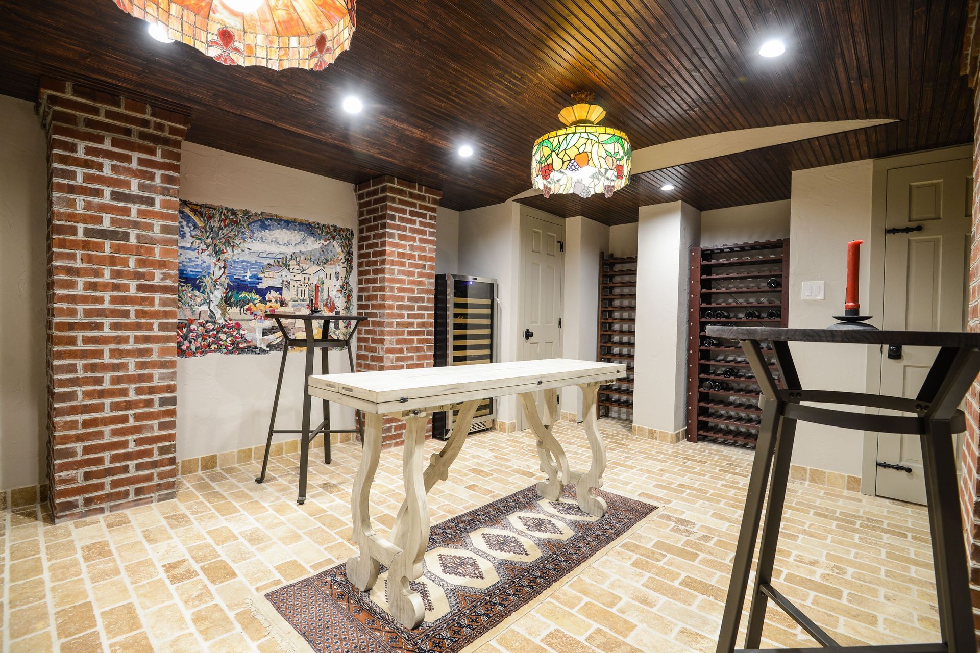 A wine cellar with a white table, two high chairs, brick pillars, a wood ceiling, and two ornate glass pendant lights.