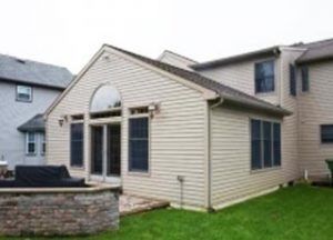 A tan vinyl-sided house addition featuring a large arched window above a sliding glass door and a small stone patio.