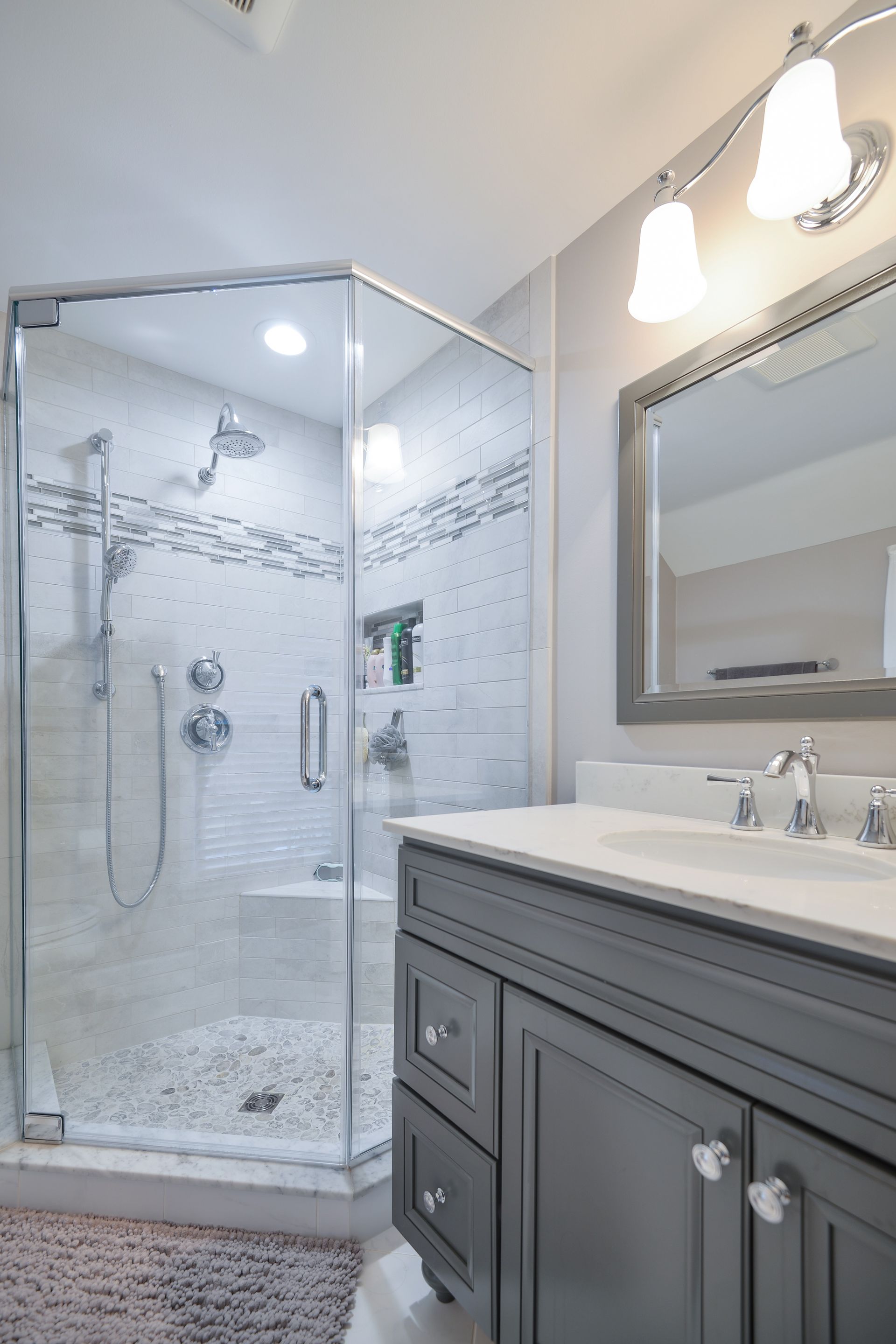A bathroom with a corner glass shower, grey vanity cabinet, white countertop, and a mirror above the sink.