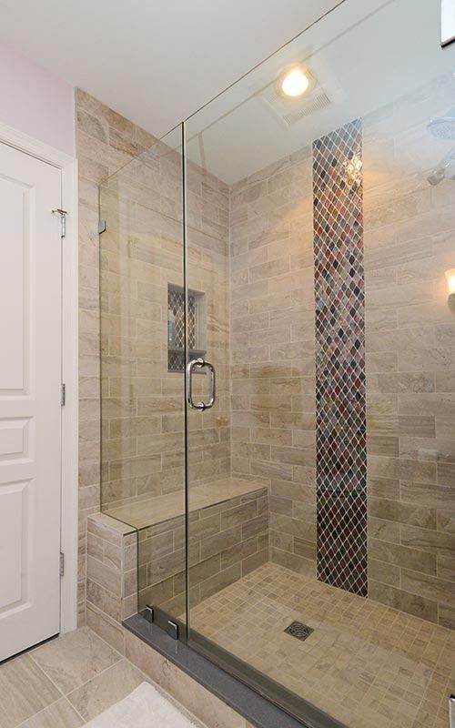 Walk-in shower with beige stone tiles, a built-in bench, glass door, and a vertical stripe of mosaic accent tiles.