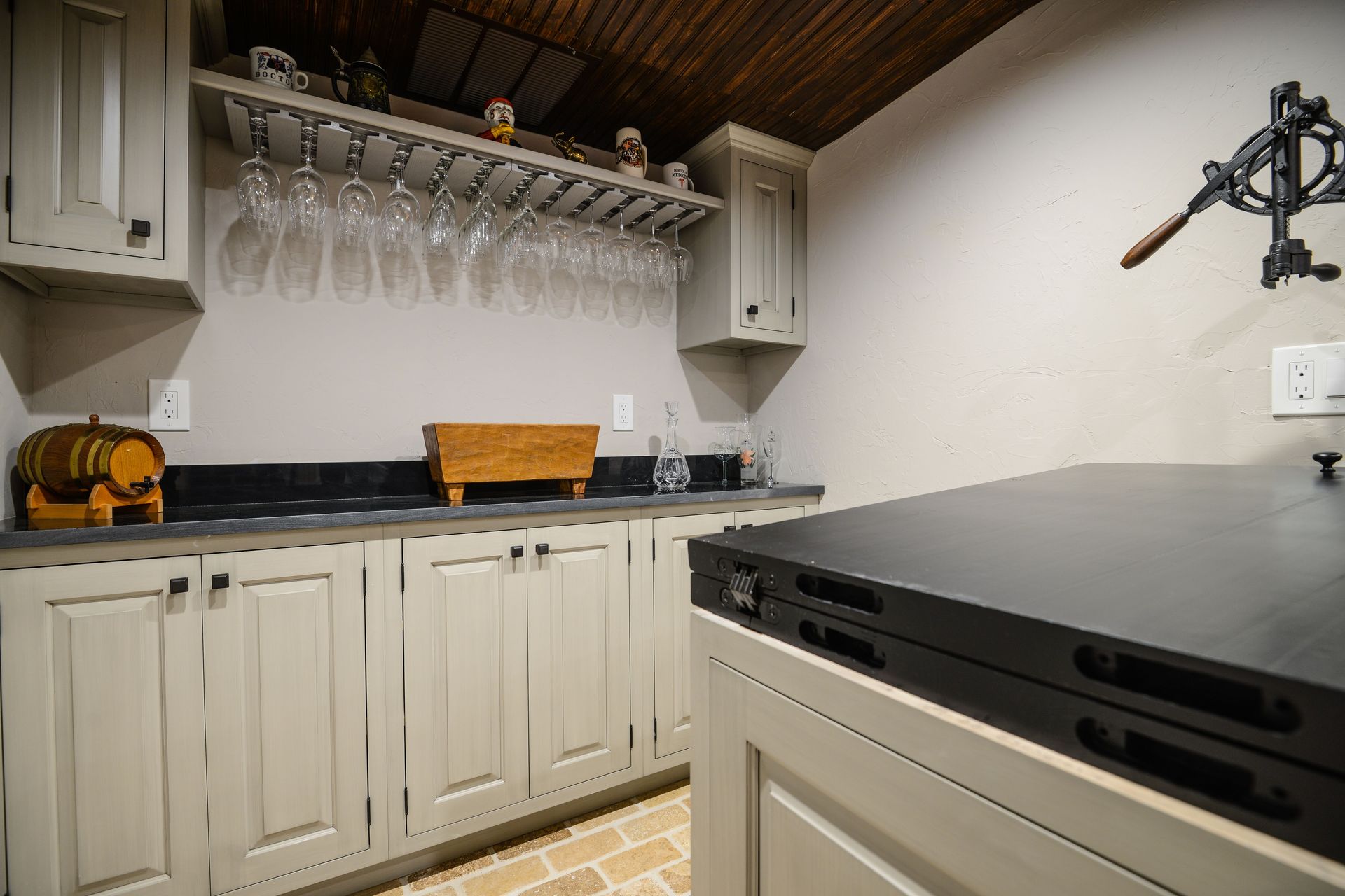 A rustic home bar with cream cabinets, a black countertop, wine glass racks, and a barrel accent under a wood ceiling.