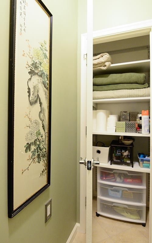 A green wall with a framed floral print next to an open closet filled with shelves of folded towels and plastic bins.