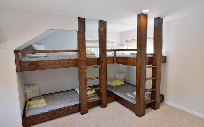 A custom wooden L-shaped bunk bed unit in a bright, minimalist room with built-in ladders and gray bedding.