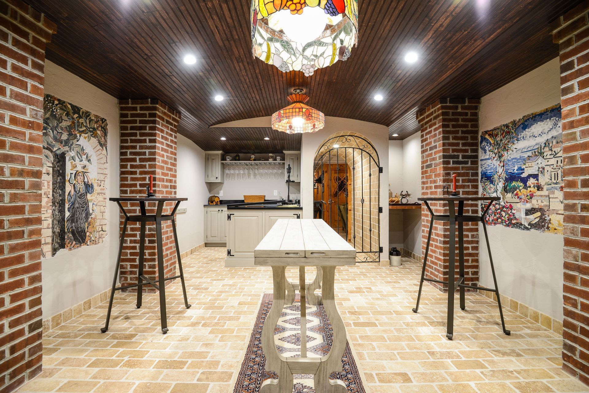 A basement wine cellar with brick columns, tiled floors, a high table, and arched entryway with decorative lighting.