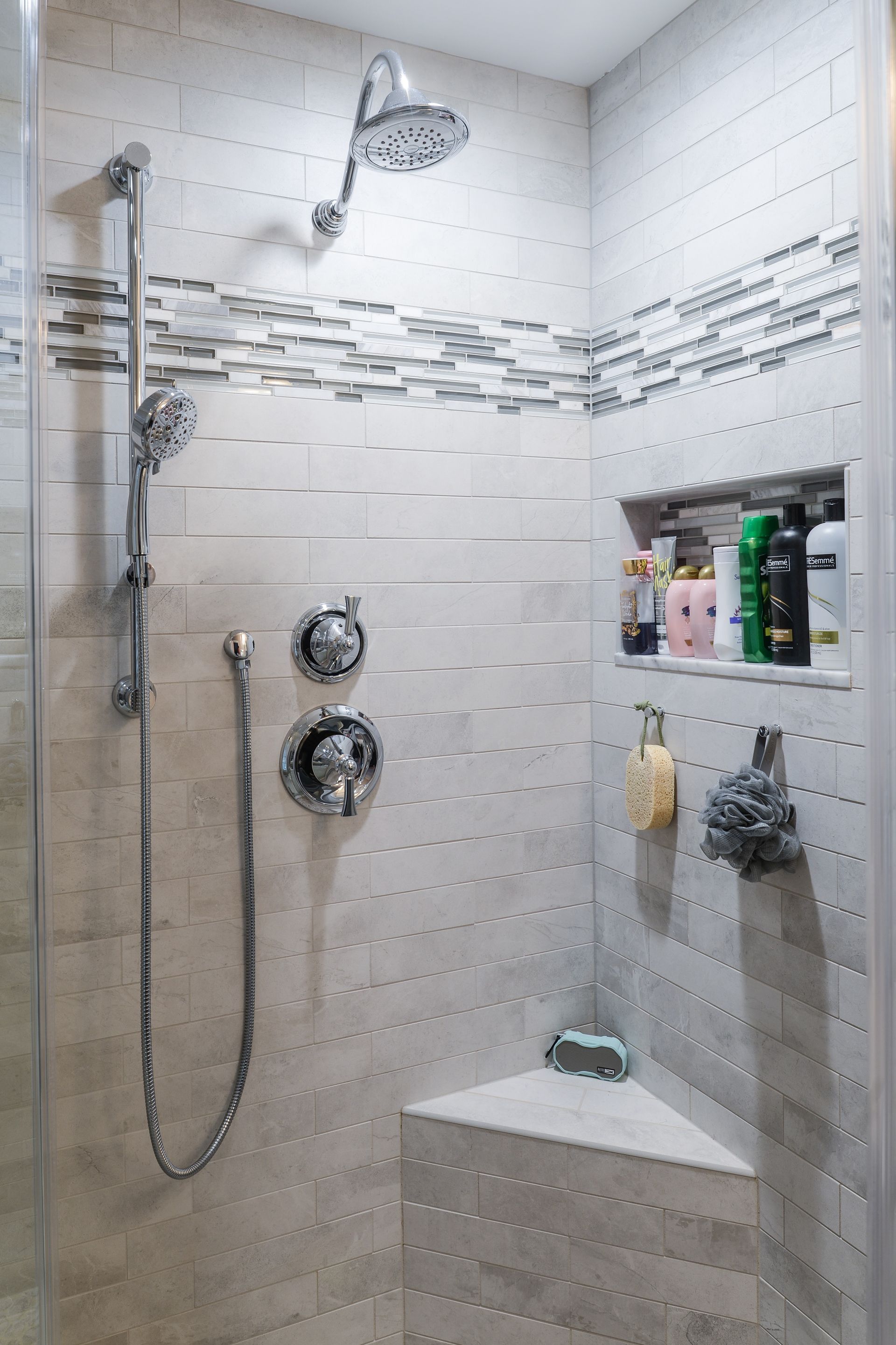 A modern shower with light-colored tile walls, a rain showerhead, handheld sprayer, corner seat, and built-in shelving.