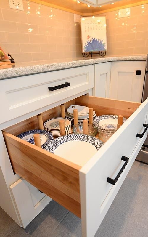 A kitchen drawer with white cabinets and black handles is open, revealing stacks of plates organized by wooden pegs.