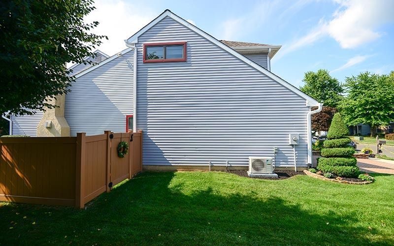 A light-blue house with gray siding, a tan fence, a manicured spiral bush, and an AC unit in a sunny, grassy yard.