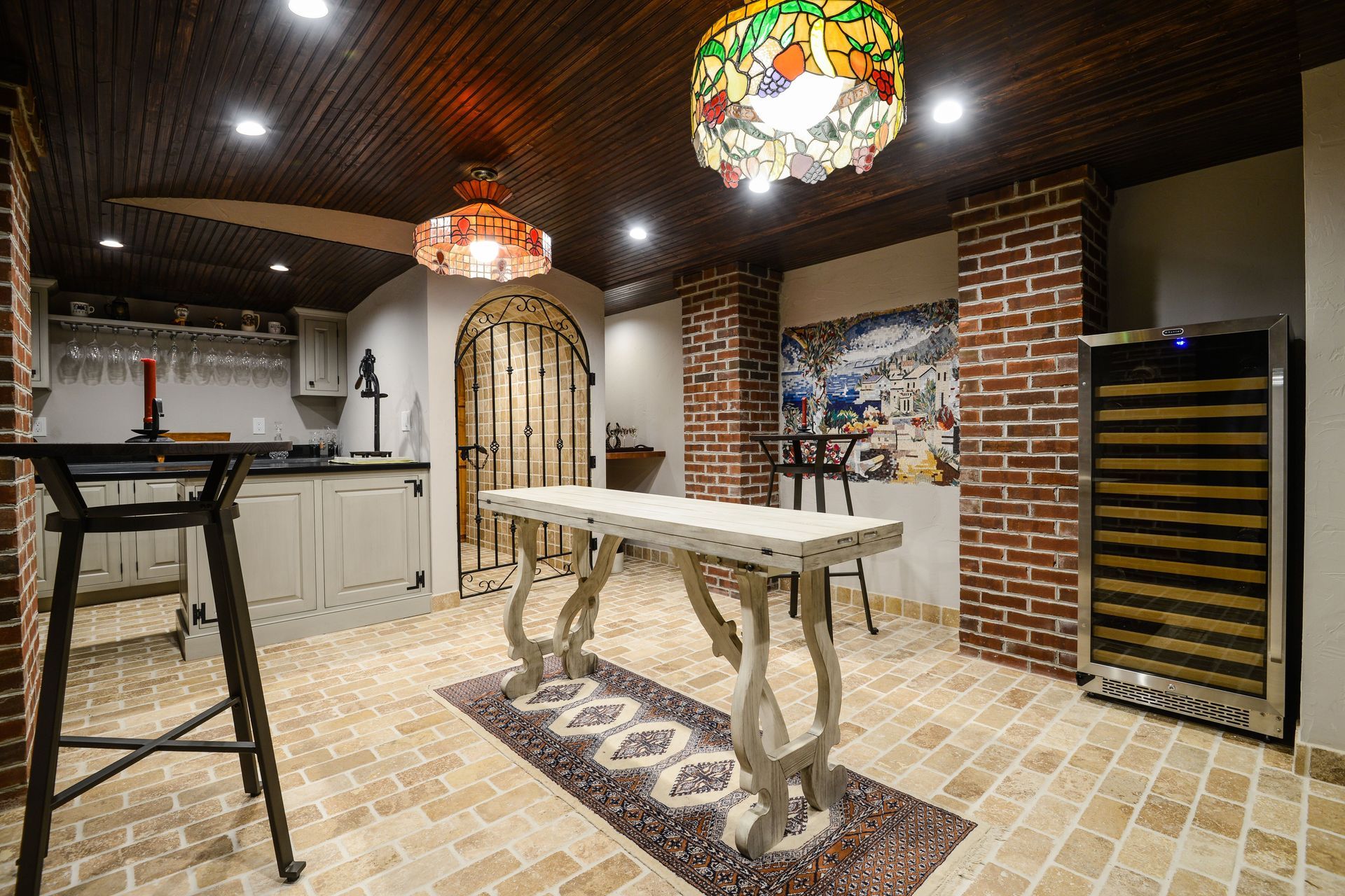 A wine cellar featuring a long table, tall stools, brick pillars, wood-paneled ceiling, and a built-in wine refrigerator.