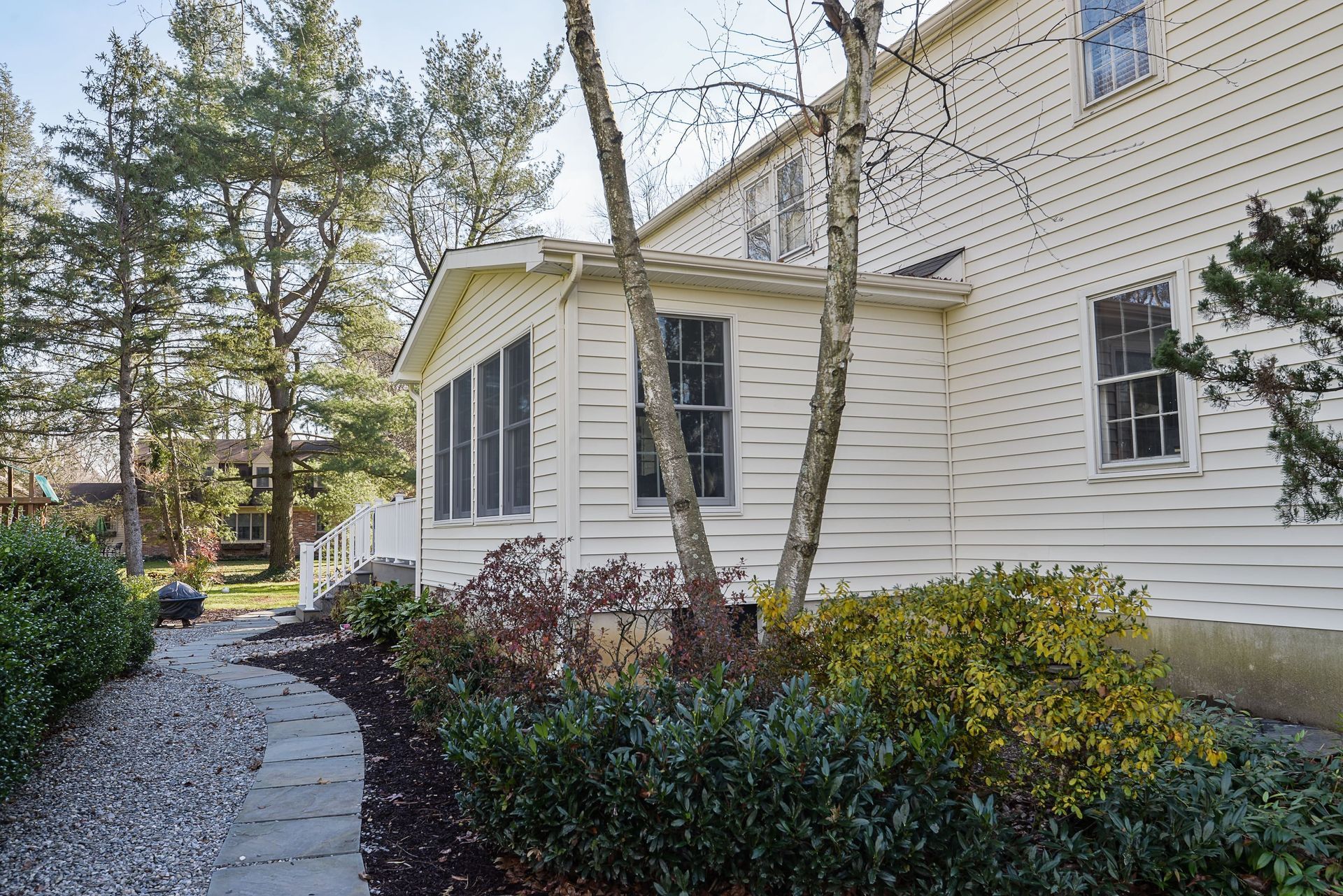 A flagstone path leads through a garden toward the side of a light-colored house with a screen porch and mature trees.