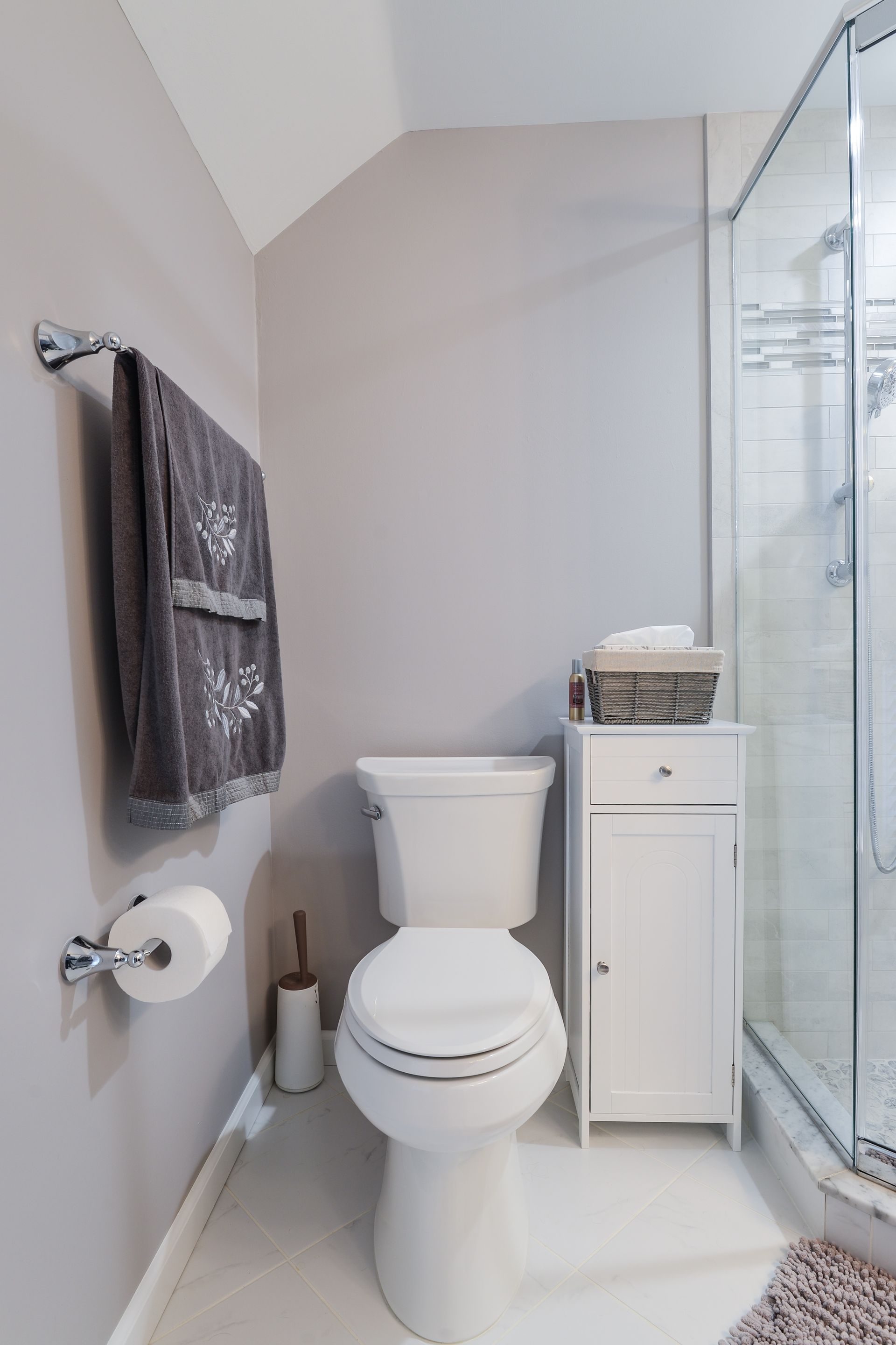 A white toilet and a thin white storage cabinet sit between a grey wall with a towel and a glass-enclosed shower.
