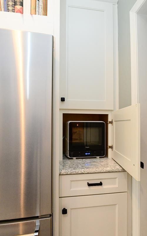A small microwave sits on a granite countertop inside an open cabinet next to a stainless steel refrigerator.