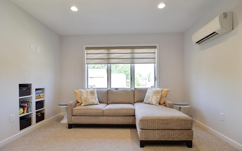 A cozy living room with a beige sectional sofa in front of a window, built-in shelving, and a wall-mounted air conditioner.