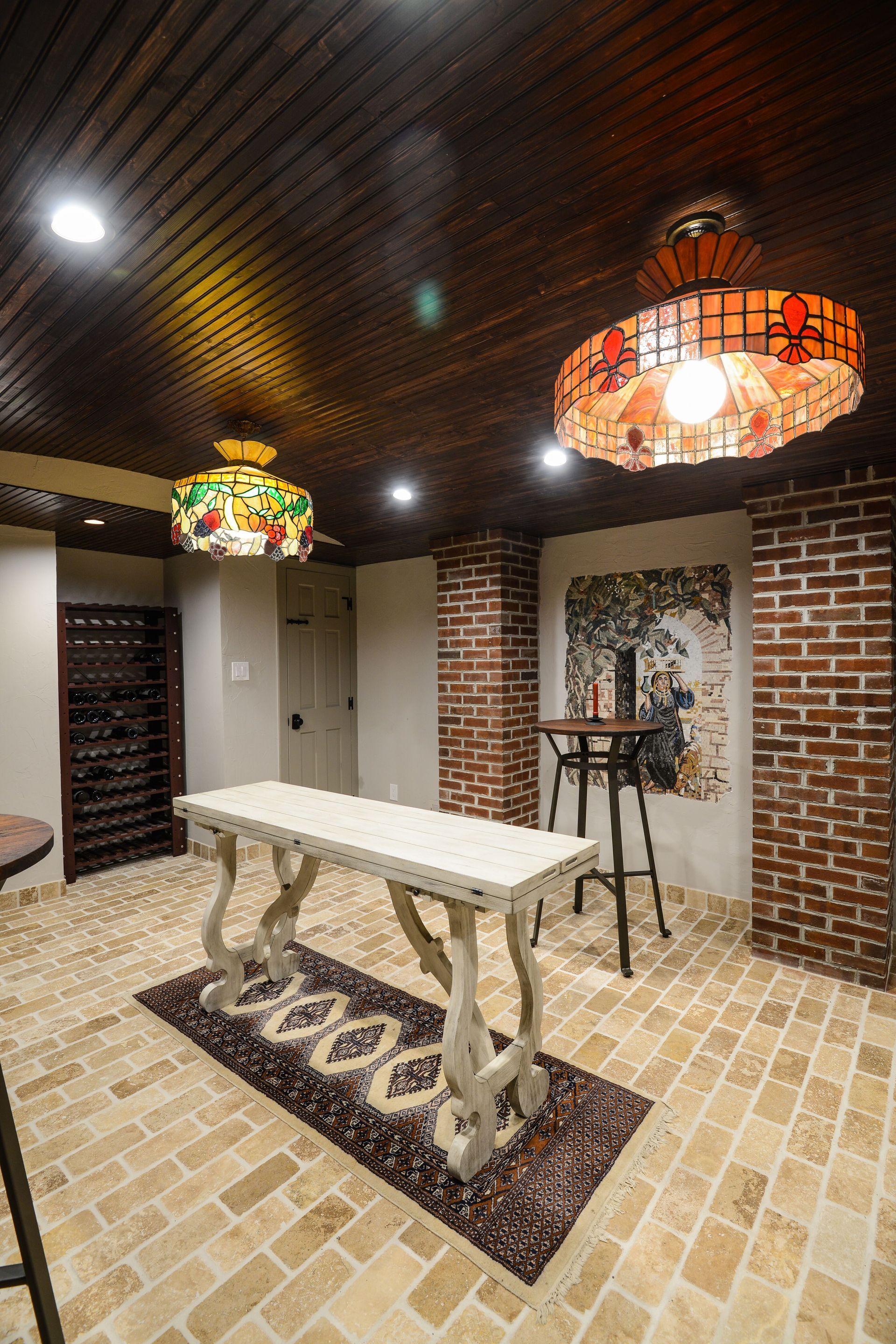 A rustic wine cellar features a long white table on a rug, a wooden ceiling, two stained-glass lamps, and brick columns.