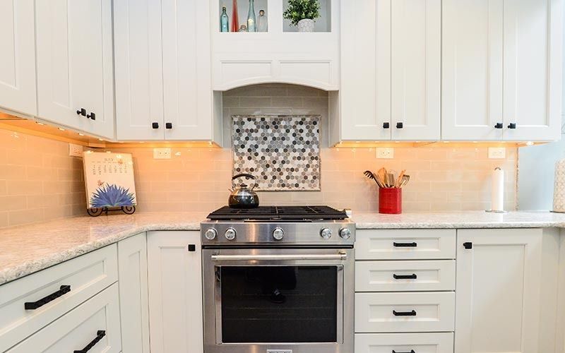 White kitchen cabinets surround a stainless steel gas range with a mosaic backsplash and under-cabinet lighting.