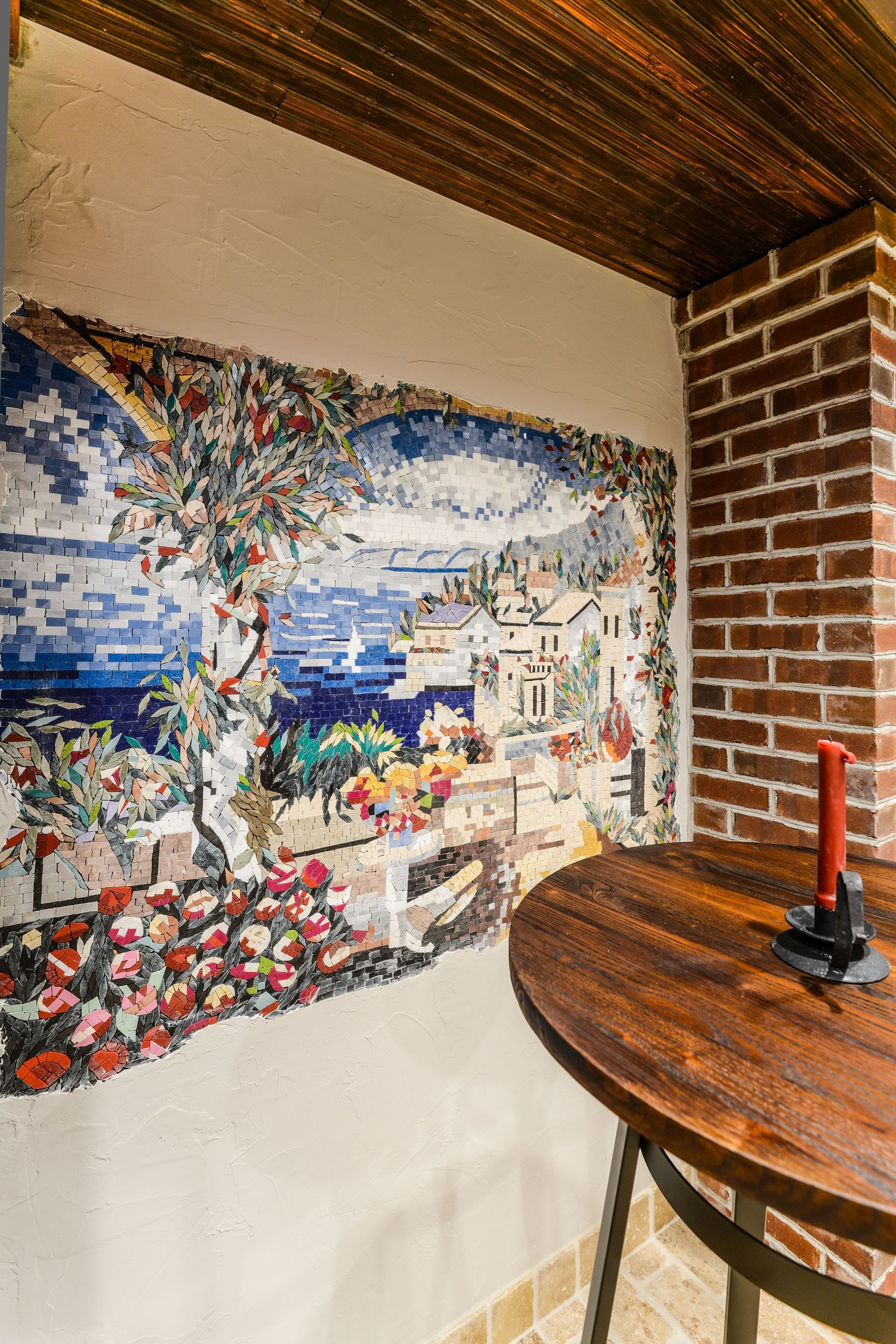 A large Mediterranean-style mosaic mural on a textured wall next to a brick pillar and a round wooden table.