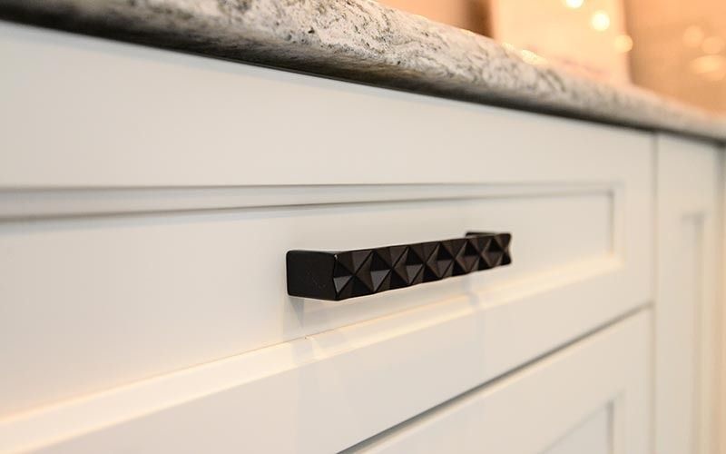 A textured matte black drawer pull centered on a white, paneled cabinet door with a gray speckled granite countertop.
