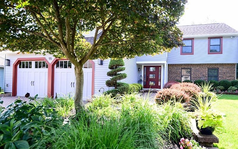 A two-story suburban home with a two-car garage, brick and light-colored siding, and a lush front garden with a large tree.