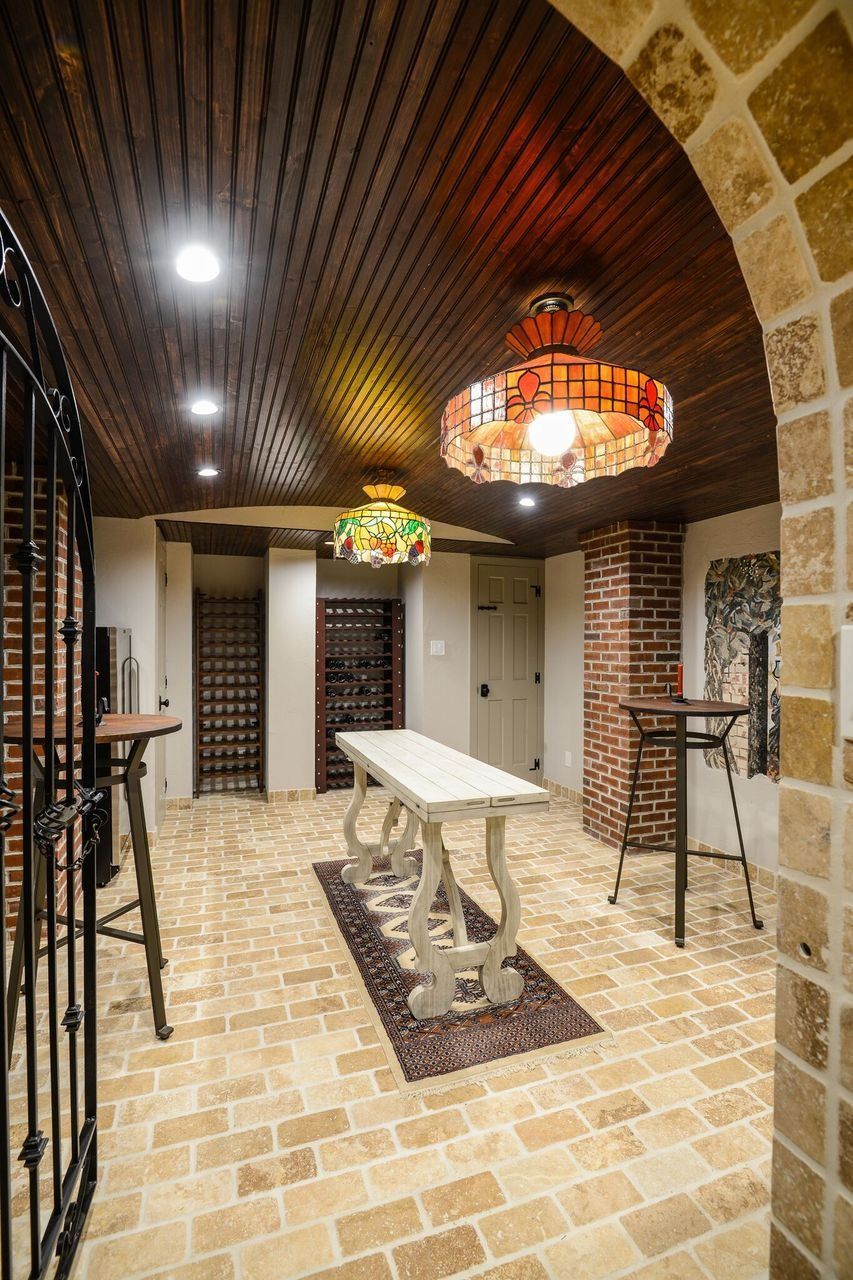 A rustic wine cellar with arched wood-slat ceiling, tan brick flooring, two light fixtures, and a central white table.