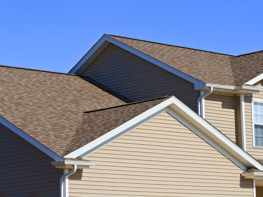 Tan vinyl siding and brown shingled roofs of a multi-level suburban home under a clear blue sky.