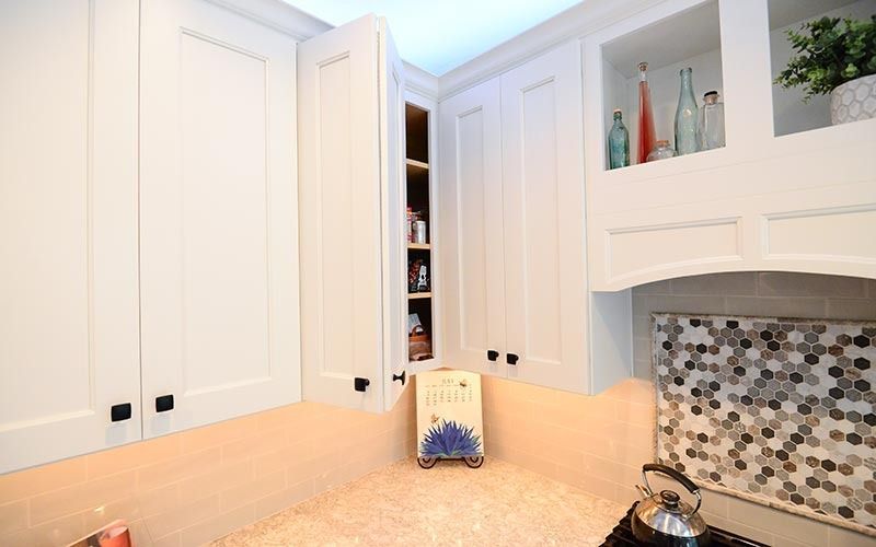 A kitchen counter with white cabinets, a small open shelf, a mosaic tile backsplash, and decorative bottles above a stove.
