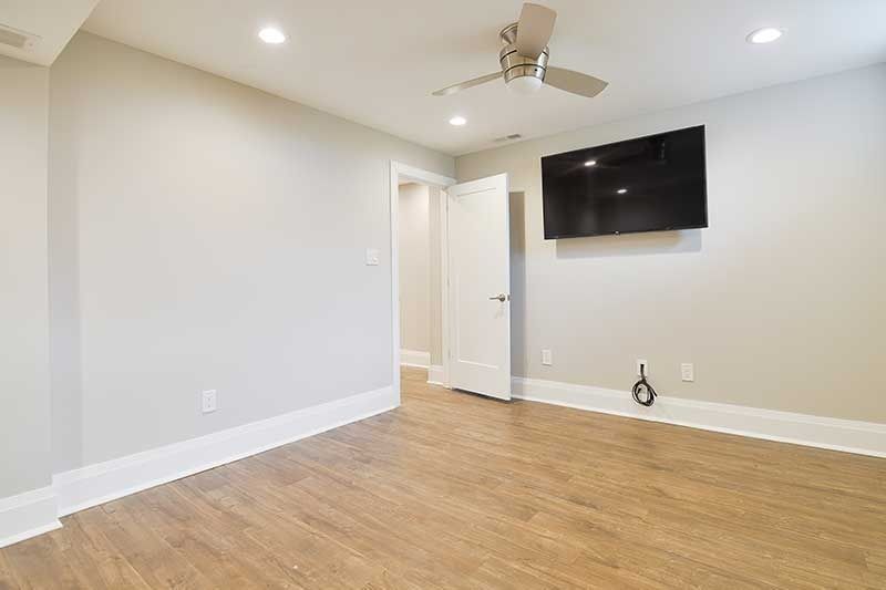 A modern bedroom with light gray walls, wood-look flooring, a wall-mounted TV, and a white door leading to another room.