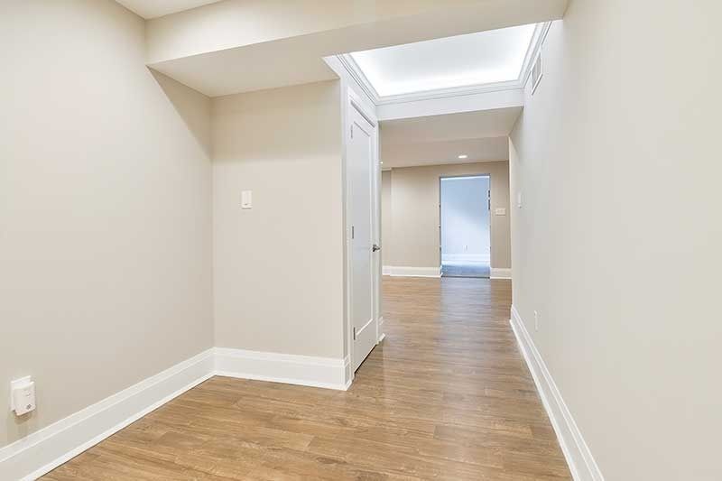 A hallway with light wood floors and beige walls, leading to an open doorway with a view into another bright room.