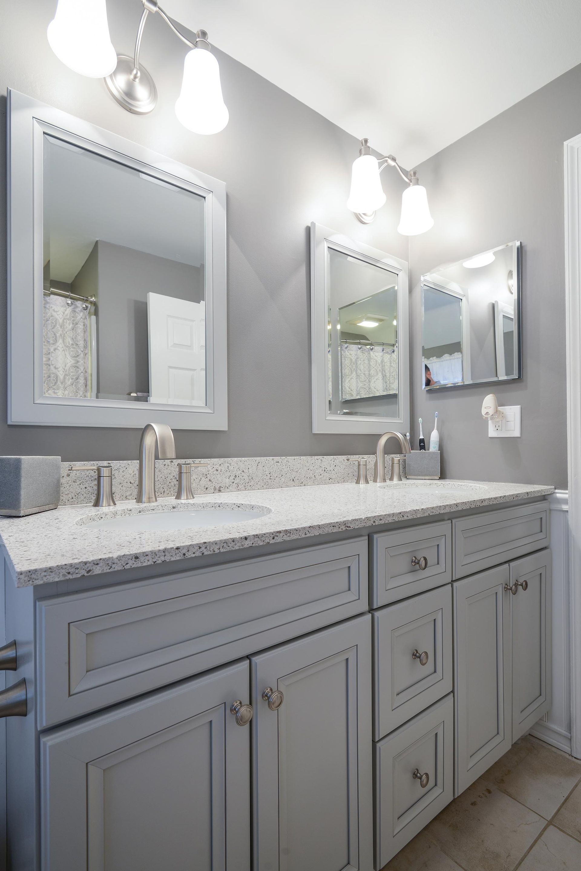 Double vanity with grey cabinets, light speckled stone countertop, two rectangular mirrors, and silver lighting fixtures.