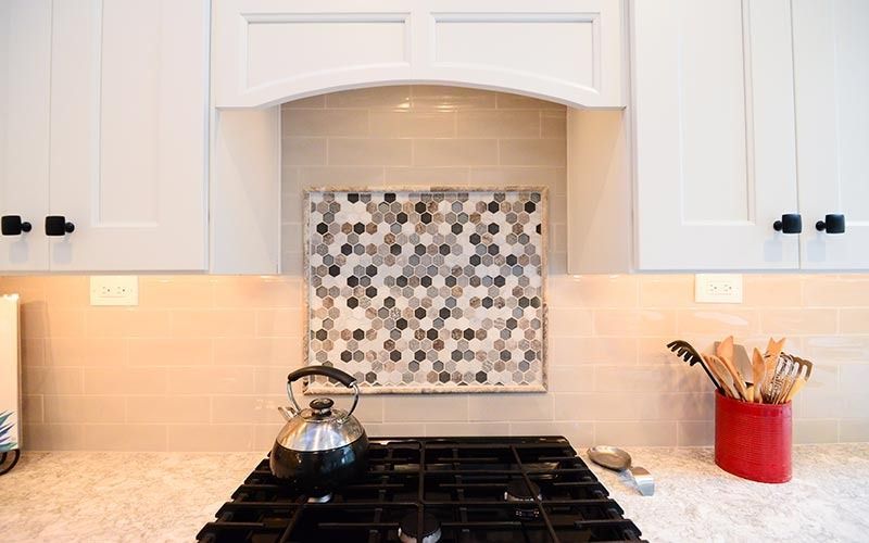 A kitchen stovetop with a patterned hexagonal tile backsplash, white cabinets, and a red utensil holder.