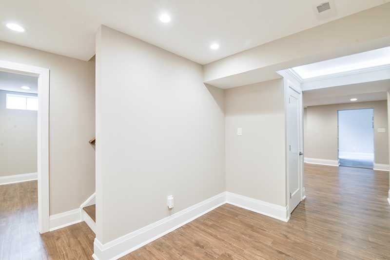 A modern basement hallway with light grey walls, white trim, and wood-look flooring, showing doorways into other rooms.
