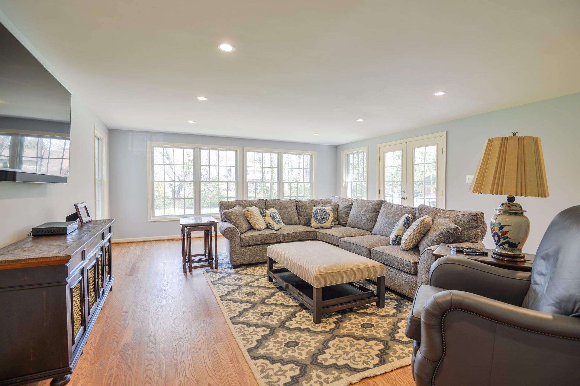 A living room with a grey sectional, a patterned rug, a wooden coffee table, a TV console, and a lamp on a table.