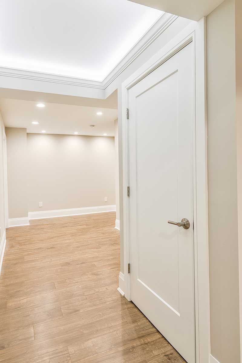 A white door stands in the foreground of a hallway with light-colored flooring and walls, lit by recessed ceiling lights.