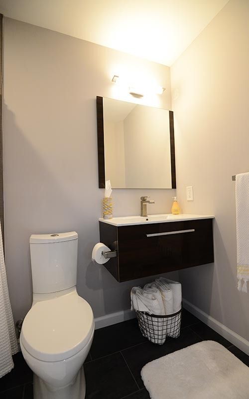 A modern bathroom featuring a white toilet, a floating dark wood vanity with a sink, a wall mirror, and a towel basket.