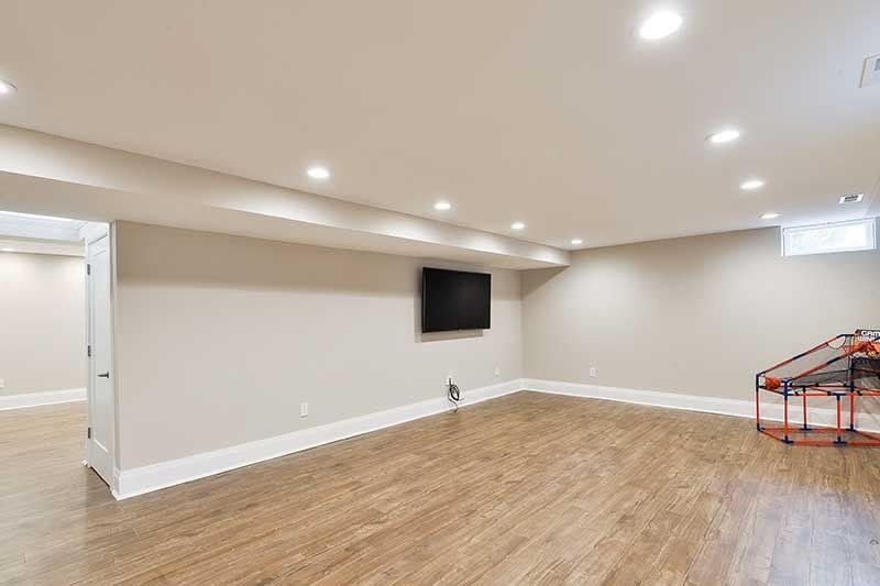 A spacious basement with light-colored walls, wood-look flooring, recessed lighting, a mounted TV, and a toy basketball hoop.