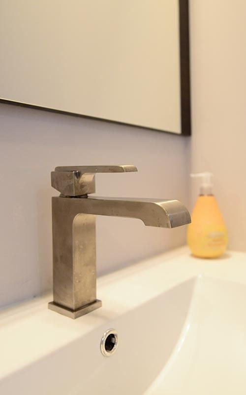 A modern, brushed-metal bathroom faucet sits on a white basin with a bottle of yellow soap nearby and a mirror above.