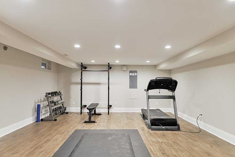 A home gym in a basement with light wood flooring, a treadmill, a weight rack, a bench, and recessed ceiling lights.